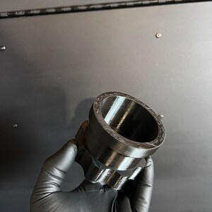 May include: A black plastic 3D printed adapter with a cylindrical shape and a textured surface.