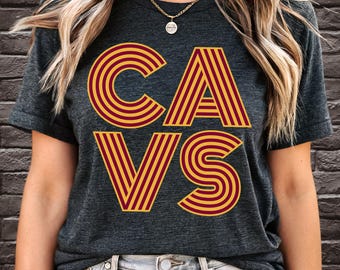 Pode incluir: Uma camiseta cinza escura com um gráfico de estilo retrô da palavra "CAVS" em listras amarelas e vinho.