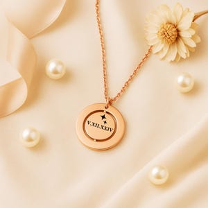 May include: Rose gold pendant necklace with a circular design, featuring the Roman numerals V.XII.XXIV and two star accents. The necklace is displayed with pearls and a flower on a cream-colored fabric.