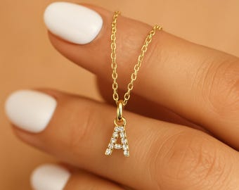May include: A gold-coloured necklace featuring a pendant in the shape of the letter "A". The pendant is adorned with small, clear gemstones. The necklace is held by a person with white nail polish.