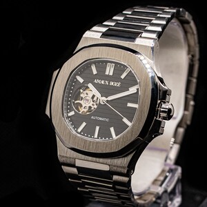 Op de afbeelding: Een zilveren horloge met een zwarte wijzerplaat en een zilveren metalen band. Het horloge heeft een skeletmechanisme en de tekst "AMAUX BOZE" en "AUTOMATIC" op de wijzerplaat.