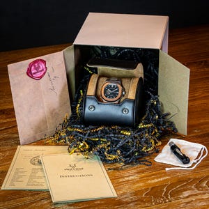 Op de afbeelding: Een bruine houten doos met een zwarte leren horlogehoes erin. De horlogehoes bevat een zilveren en zwarte polshorloge. De doos is gevuld met zwart en goud geribbeld papier. Er is een witte zak met een zwart gereedschap erin en een boekje met de tekst "Amax Boze Instructions" op de omslag.