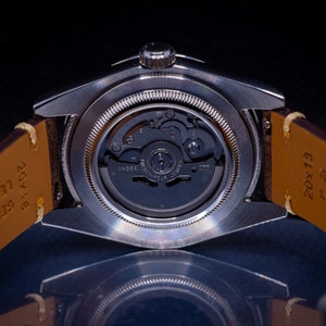 May include: Close-up of a silver wristwatch with a transparent back, revealing the intricate inner workings of the watch mechanism. The watch has a brown leather strap. The watch face is not visible.