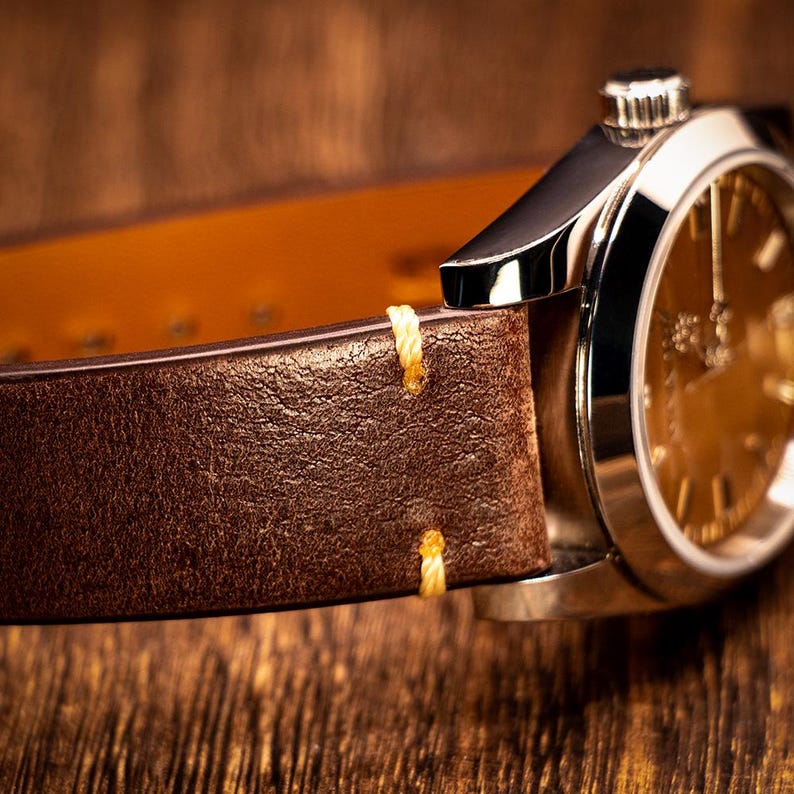 May include: Close-up of a luxury wristwatch with a brown leather strap. The watch face is gold-toned, and the strap is stitched with yellow thread. The watch case is silver-colored, and the background is a wooden surface.