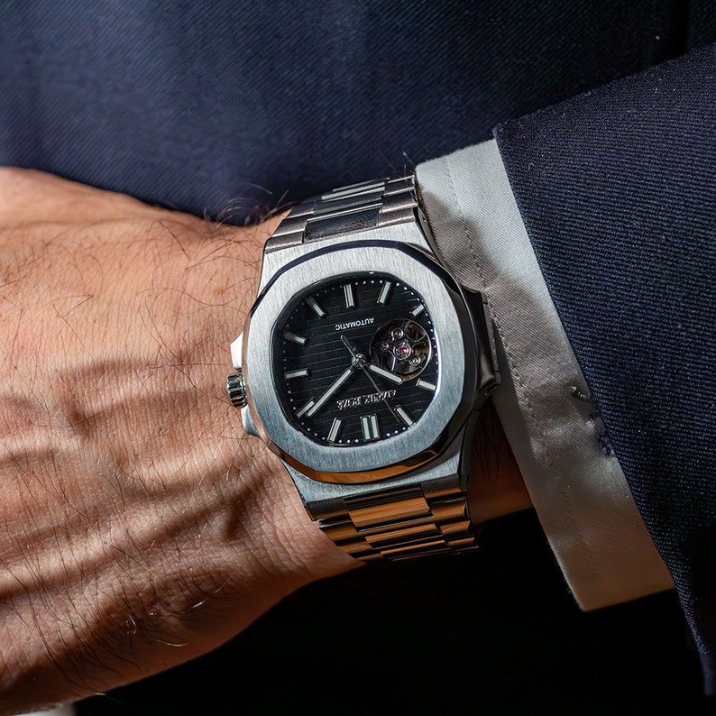 Op de afbeelding: Een zilveren polshorloge met een zwarte wijzerplaat en een skeletmechanisme. Het horloge heeft een vierkante kast en een metalen band.