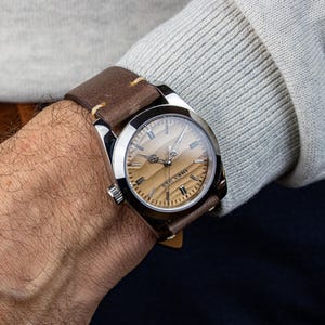 May include: A wristwatch with a brown leather band is displayed on a wrist. The watch has a silver-colored case and a light brown face with silver hands and markers. The band is stitched with cream-colored thread.