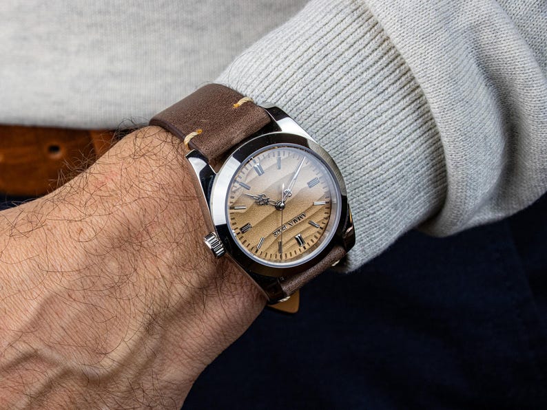 May include: A wristwatch with a brown leather band is displayed on a wrist. The watch has a silver-colored case and a light brown face with silver hands and markers. The band is stitched with cream-colored thread.
