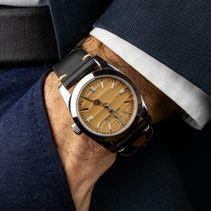 May include: A wristwatch with a silver case and a light brown face. The watch has black leather straps and is being worn on a wrist. The watch face has gold-colored markers and hands. The watch is part of a formal outfit.
