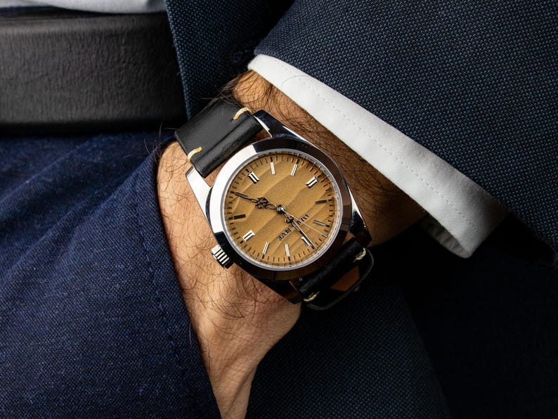 May include: A wristwatch with a silver case and a light brown face. The watch has black leather straps and is being worn on a wrist. The watch face has gold-colored markers and hands. The watch is part of a formal outfit.