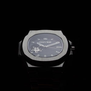 Op de afbeelding: Een zilveren polshorloge met een zwarte wijzerplaat en een skeletmechanisme. De wijzerplaat heeft witte markeringen en de tekst "AMAUX BOZ&Eacute;" in wit.