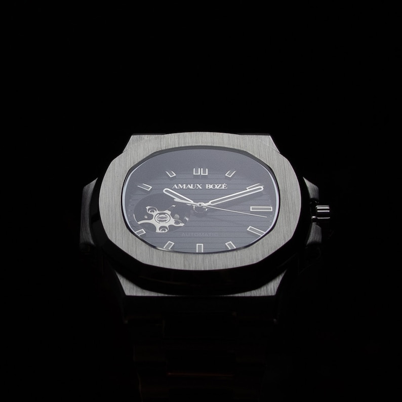 Op de afbeelding: Een zilveren polshorloge met een zwarte wijzerplaat en een skeletmechanisme. De wijzerplaat heeft witte markeringen en de tekst "AMAUX BOZ&Eacute;" in wit.