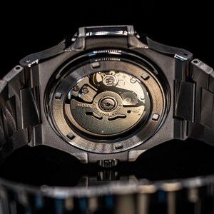 Op de afbeelding: Een zwart stalen horloge met een zilveren wijzerplaat en een zichtbaar automatisch uurwerk. Het horloge heeft een doorzichtige achterkant die de innerlijke werking van het horloge laat zien.