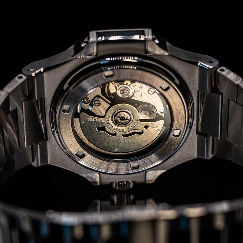 Op de afbeelding: Een zwart stalen horloge met een zilveren wijzerplaat en een zichtbaar automatisch uurwerk. Het horloge heeft een doorzichtige achterkant die de innerlijke werking van het horloge laat zien.