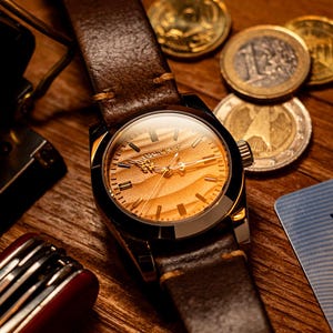 May include: A close-up of a wristwatch with a brown leather band and a gold-toned face. The watch face has a unique wood grain pattern. The watch is set against a wooden surface with coins and a small red and silver tool.