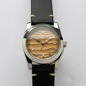 May include: A silver wristwatch with a black leather band and a tan face. The watch face has a textured design and the brand name "AMALX BOZE" is printed on the face. The band is stitched with yellow thread.