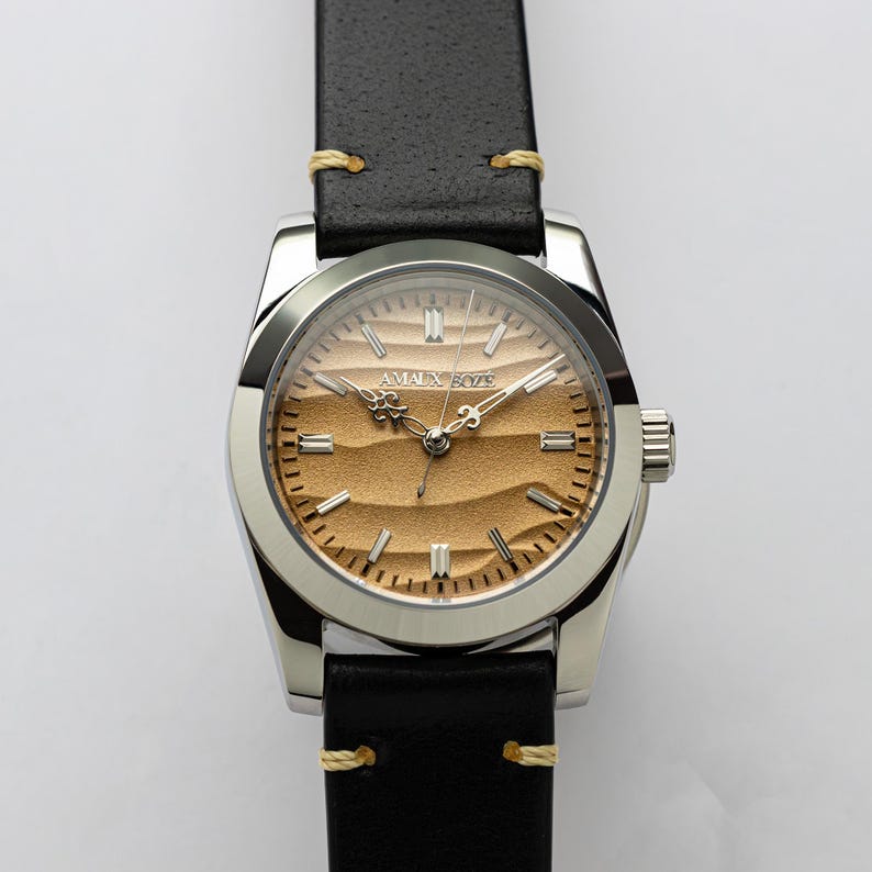 May include: A silver wristwatch with a black leather band and a tan face. The watch face has a textured design and the brand name "AMALX BOZE" is printed on the face. The band is stitched with yellow thread.