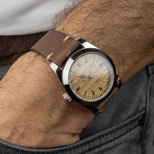 May include: A wristwatch with a light brown wooden face and silver casing. The watch has a brown leather band with cream-colored stitching. The watch is on a wrist with a gray denim pocket.