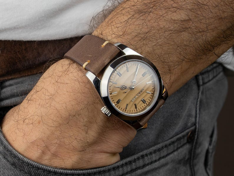 May include: A wristwatch with a light brown wooden face and silver casing. The watch has a brown leather band with cream-colored stitching. The watch is on a wrist with a gray denim pocket.