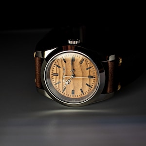 May include: A close-up of a wristwatch with a light brown wooden face and a silver-colored case. The watch has black hour markers and hands. The watchband is brown leather with visible stitching. The watch face reads "ALVIN BOAZ".