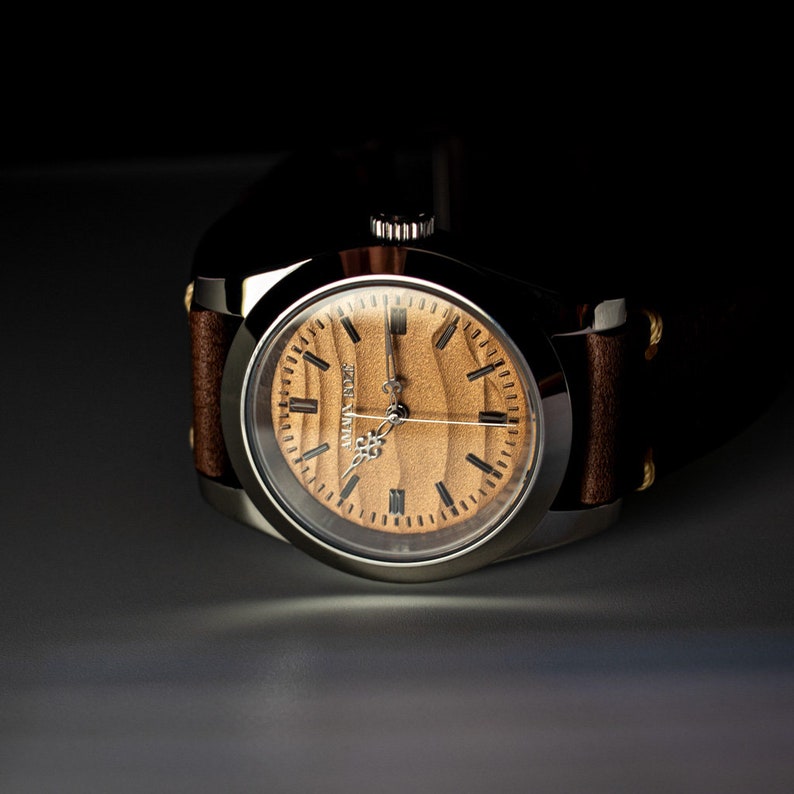May include: A close-up of a wristwatch with a light brown wooden face and a silver-colored case. The watch has black hour markers and hands. The watchband is brown leather with visible stitching. The watch face reads "ALVIN BOAZ".