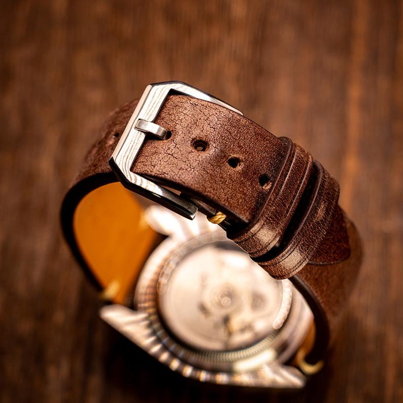 May include: Close-up of a brown leather watch strap with a silver buckle. The watch face is visible, showcasing the intricate internal mechanisms. The strap is made of textured leather, and the watch is set against a wooden background.