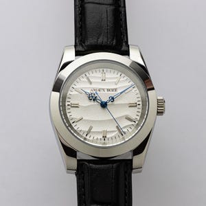 May include: A silver-toned wristwatch with a black leather band. The watch face is white with a textured wave pattern and blue hands. The brand name "AMAUX BOZE" is printed on the watch face.