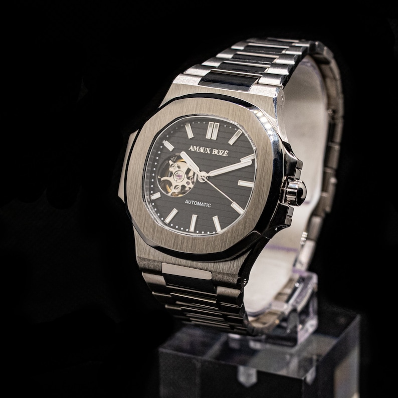 Op de afbeelding: Een zilveren horloge met een zwarte wijzerplaat en een zilveren metalen band. Het horloge heeft een skeletmechanisme en de tekst "AMAUX BOZE" en "AUTOMATIC" op de wijzerplaat.