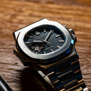 Op de afbeelding: Een zilveren polshorloge met een zwarte wijzerplaat en een zilveren band. Het horloge heeft een skeletmechanisme en de tekst "A.L.S.U.N.N. BULE" is zichtbaar op de wijzerplaat.