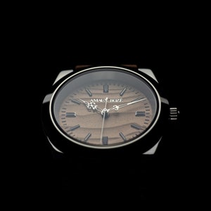 May include: A wristwatch with a brown wooden-style face and a silver-colored case. The watch has a brown leather band and the text "AMAUX BOZE" is printed on the watch face. The watch hands are silver and the background is black.