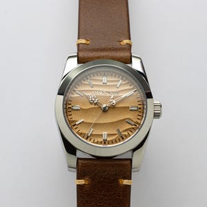 May include: A stainless steel wristwatch with a brown leather band. The watch face has a textured, sand-colored design with the text "AMAL X 5076". The band is secured with light brown stitching.