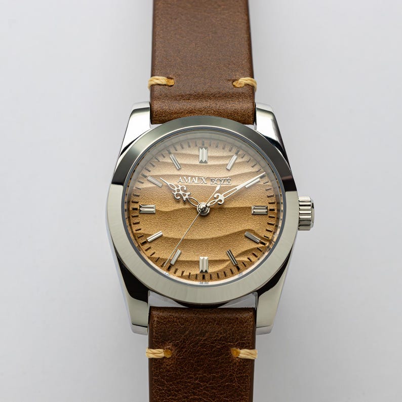 May include: A stainless steel wristwatch with a brown leather band. The watch face has a textured, sand-colored design with the text "AMAL X 5076". The band is secured with light brown stitching.