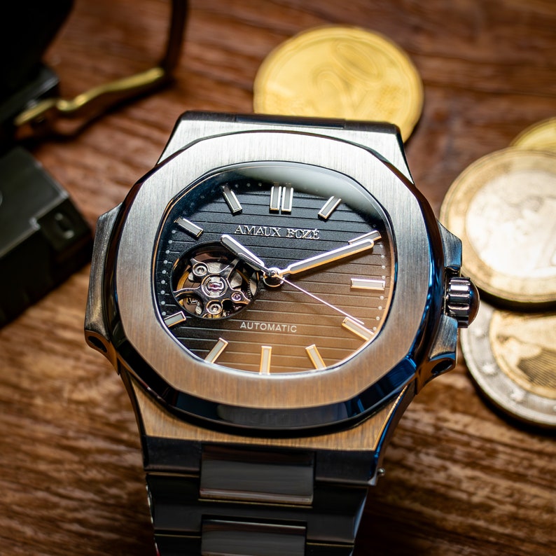 Op de afbeelding: Een zilveren polshorloge met een zwart en bruin wijzerplaat. Het horloge heeft een skeletmechanisme en de tekst "Amauix Boxe Automatic" op de wijzerplaat.