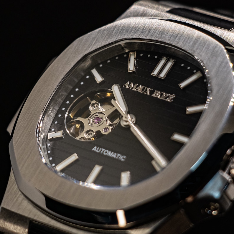 Op de afbeelding: Een zilveren horloge met een zwarte wijzerplaat en een geskeletteerd automatisch uurwerk. De wijzerplaat heeft witte uurmarkeringen en de tekst "AMAX RYZ" bovenaan.