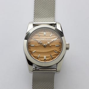 May include: A silver wristwatch with a mesh band. The watch face is a light brown color with a textured pattern and silver hands. The watch has the text "AMAUX 5076" on the face.