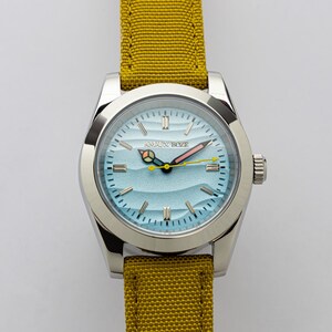 Peut inclure: Une montre-bracelet argentée avec un cadran bleu clair représentant des vagues et le texte "AMAIX BOZE". La montre a un bracelet en toile jaune. Les aiguilles sont roses et jaunes.