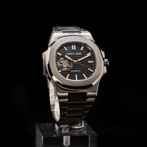 Op de afbeelding: Een zilveren polshorloge met een zwarte wijzerplaat en een zilveren band. Het horloge heeft een skeletmechanisme en de tekst "AMAUX BOXE" en "AUTOMATIC" op de wijzerplaat.