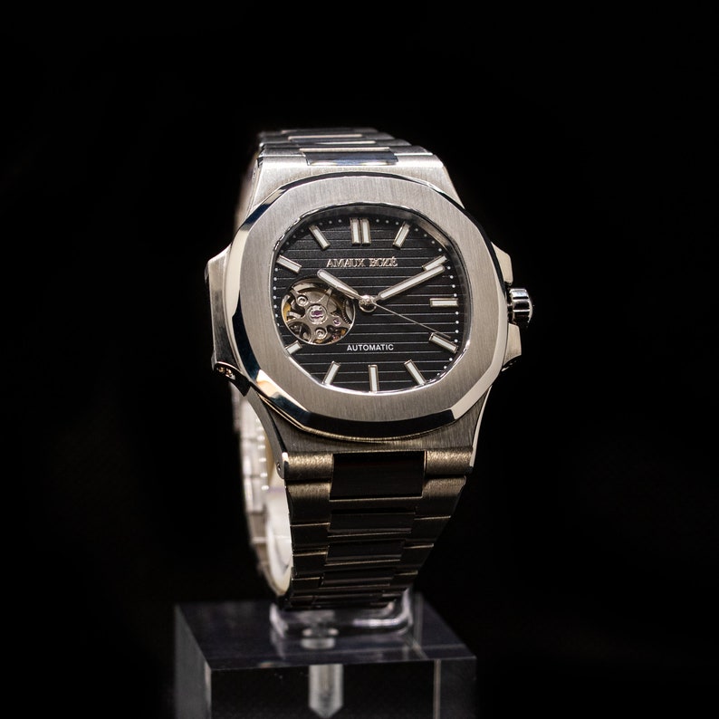 Op de afbeelding: Een zilveren polshorloge met een zwarte wijzerplaat en een zilveren band. Het horloge heeft een skeletmechanisme en de tekst "AMAUX BOXE" en "AUTOMATIC" op de wijzerplaat.