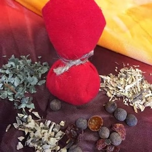 May include: A bright red fabric pouch tied with a piece of natural jute twine, surrounded by scattered dried herbs, small dark brown spherical berries, and light-colored woody plant fragments on a dark red textured surface.