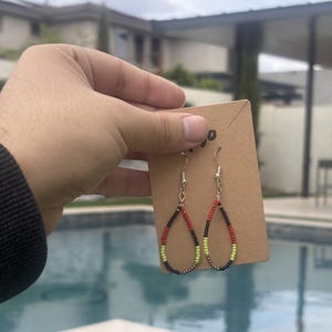 May include: Handmade teardrop earrings with a beaded design. The earrings feature a silver hook and a pattern of black, red, and yellow-green beads. The earrings are displayed on a brown card.