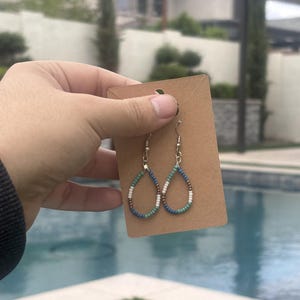 May include: Teardrop-shaped beaded earrings. The earrings feature a silver hook and are adorned with small beads in white, blue, and brown. The earrings are displayed on a brown card.