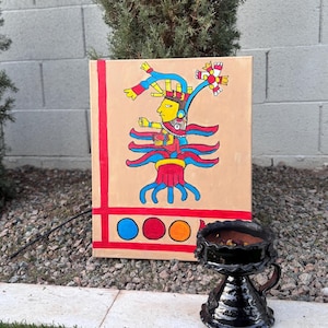 May include: An Aztec-inspired painting on canvas featuring a stylized figure with a yellow face and elaborate headdress. The artwork is framed by red lines and includes three colored circles. A black ceramic vessel with a handle sits in front of the painting.
