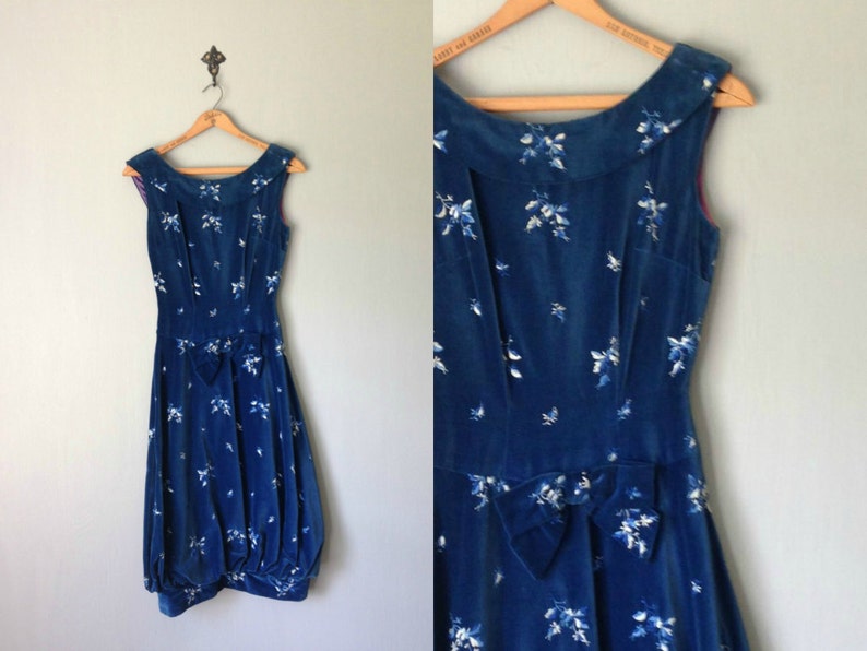 Puede incluir: Un vestido de terciopelo azul con estampado floral blanco y un lazo en la cintura. El vestido es sin mangas y tiene un escote redondo.