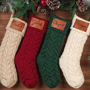 Peut inclure: Quatre bas de Noël en tricot torsadé crème, rouge et vert, chacun avec une étiquette nominative en cuir. Les noms Sophia, Jacob, Abigail et Mason sont affichés. Décorations de vacances festives avec du pin et des ornements.