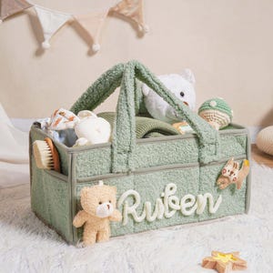 Puede incluir: Una cesta organizadora para bebés de color verde claro con el nombre "Ruben" en escritura blanca. La cesta contiene artículos para bebés, como un cepillo, juguetes y un oso de peluche. Un pequeño osito de peluche está adjunto en la parte delantera. La cesta está sobre una alfombra blanca y esponjosa.