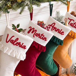 Op de afbeelding: Vier kerstsokken van fluweel met ruches en geborduurde namen. De sokken zijn wit, rood, donkergroen en mosterdgeel, met de namen Valeria, Martin, Annie en een gedeeltelijke naam, hangend met decoratieve accenten en kleine versieringen.