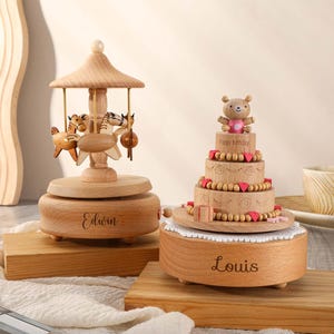 May include: Two wooden music boxes, one shaped like a carousel with horses and the other shaped like a cake with a teddy bear on top. The carousel music box is engraved with the name "Edwin" and the cake music box is engraved with the name "Louis".