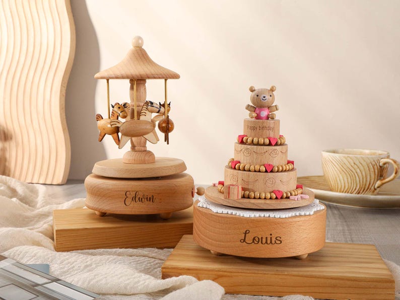 May include: Two wooden music boxes, one shaped like a carousel with horses and the other shaped like a cake with a teddy bear on top. The carousel music box is engraved with the name "Edwin" and the cake music box is engraved with the name "Louis".