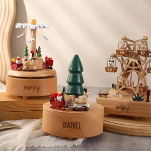 May include: Three wooden music boxes with different designs. The first music box features a carousel with a snowman and reindeer. The second music box features a Christmas tree with a reindeer and Santa Claus. The third music box features a Ferris wheel with animals. Each music box has a personalized name on the base: Henry, Daniel, and Andy.
