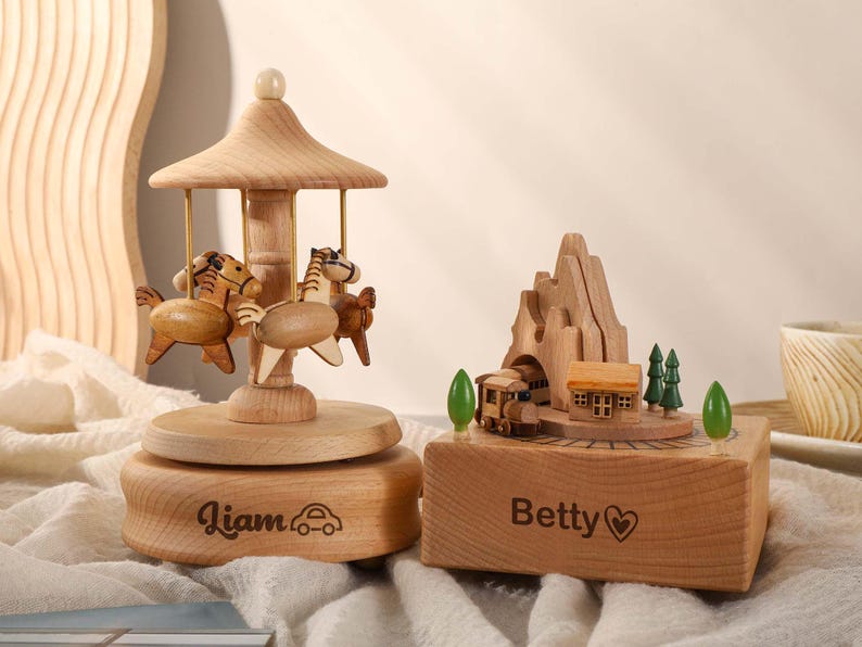 May include: Two wooden music boxes with personalized engravings. One features a carousel with horses and the name "Liam" and a car. The other features a train and a mountain scene with the name "Betty" and a heart.