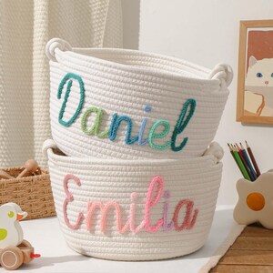 May include: Two white rope storage baskets with colourful embroidered names, "Daniel" and "Emilia". The baskets are stacked on top of each other.